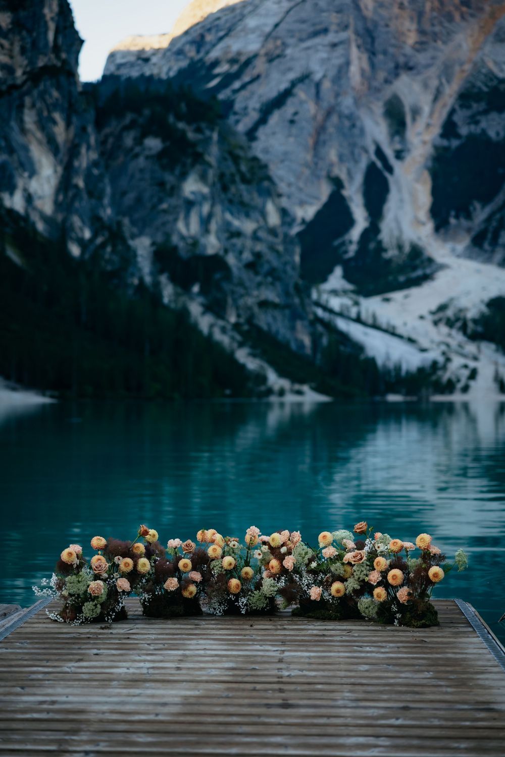 Yellow and cream floral arrangements line a wooden dock over serene blue alpine lake waters.