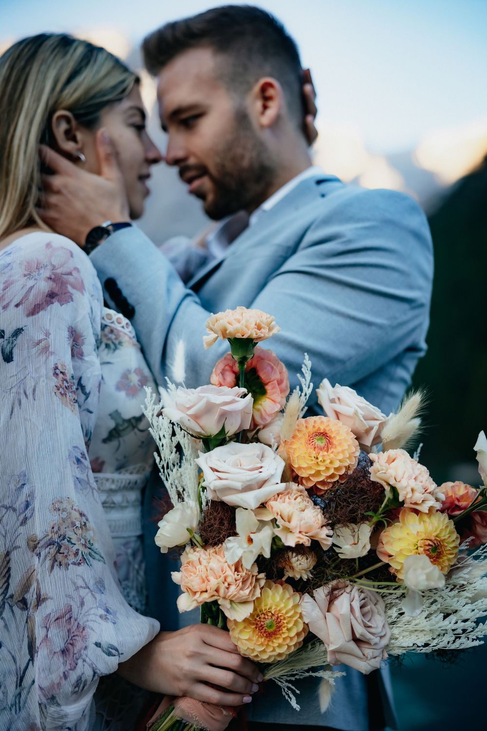 Romantic wedding couple holding peach and blush floral bouquets against mountain backdrop at sunset.