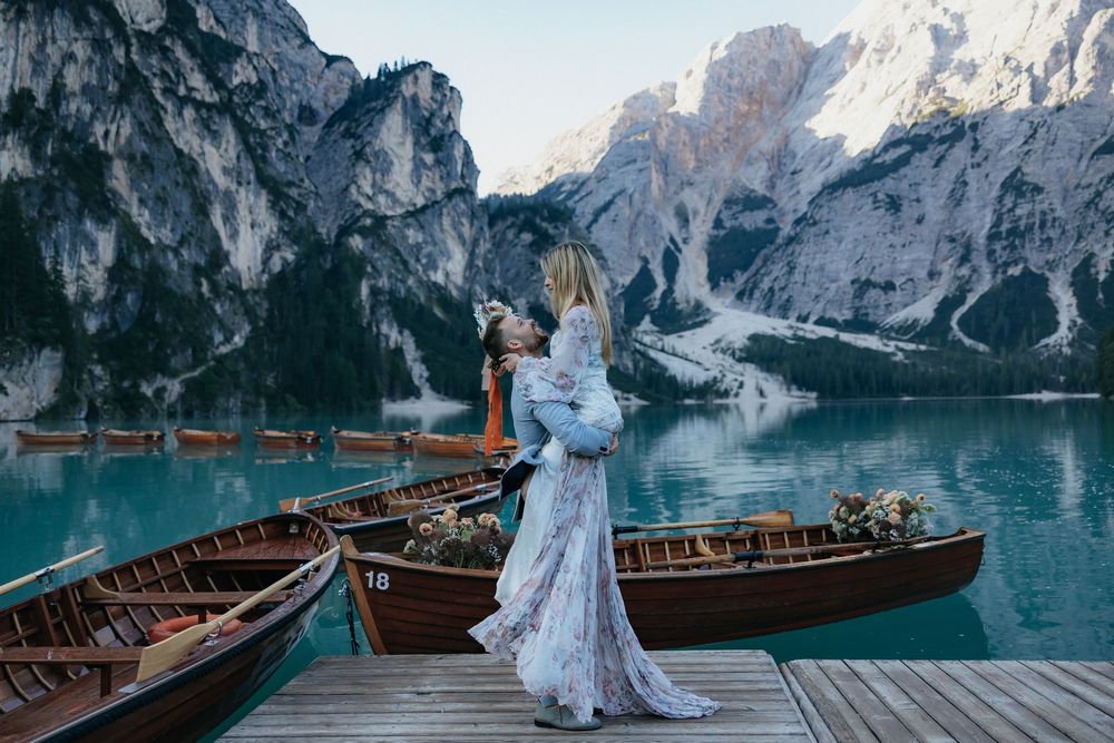 A romantic couple embraces on a wooden dock overlooking turquoise Lake Braies with Dolomite mountains in background.