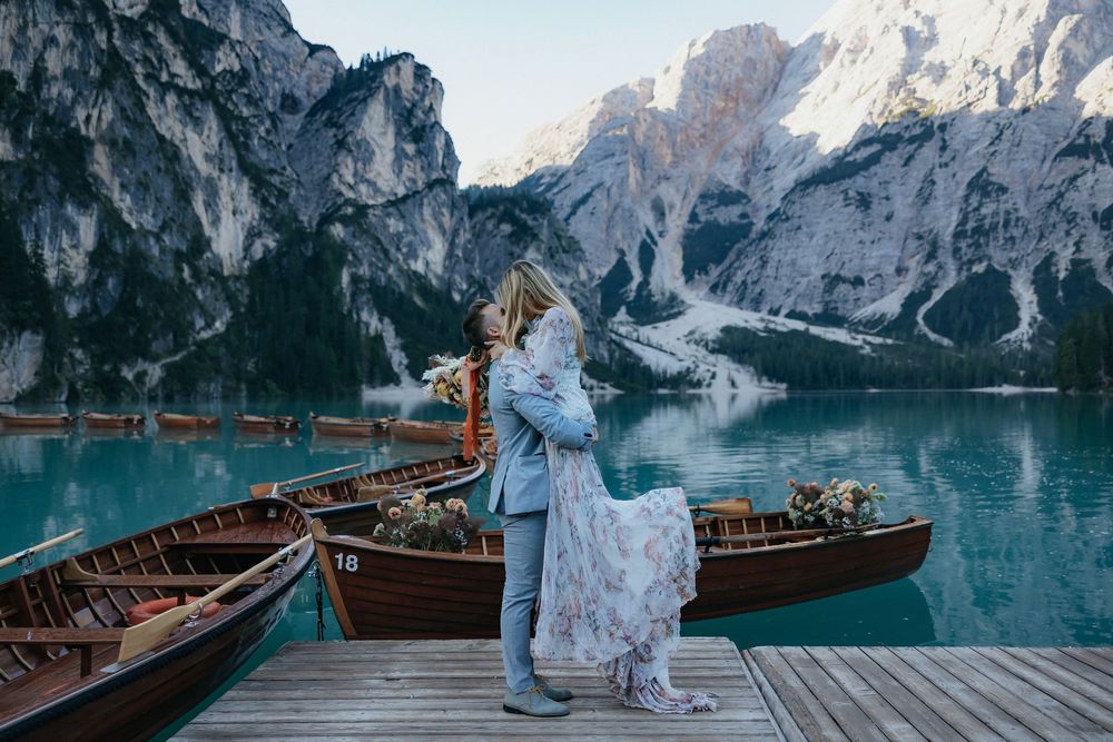 A couple in wedding attire stands on a wooden dock with traditional wooden boats against dramatic mountain peaks at Lake Braies.