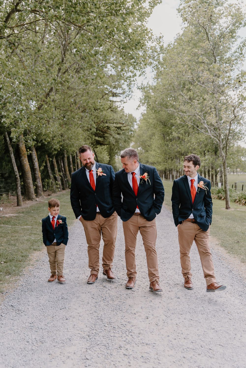 Wedding party members in navy jackets and khaki pants with red ties walk together on a tree-lined gravel path.