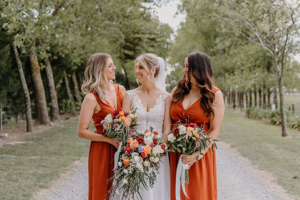 Three people in rust-colored and white dresses hold floral bouquets while walking on a tree-lined path outdoors.