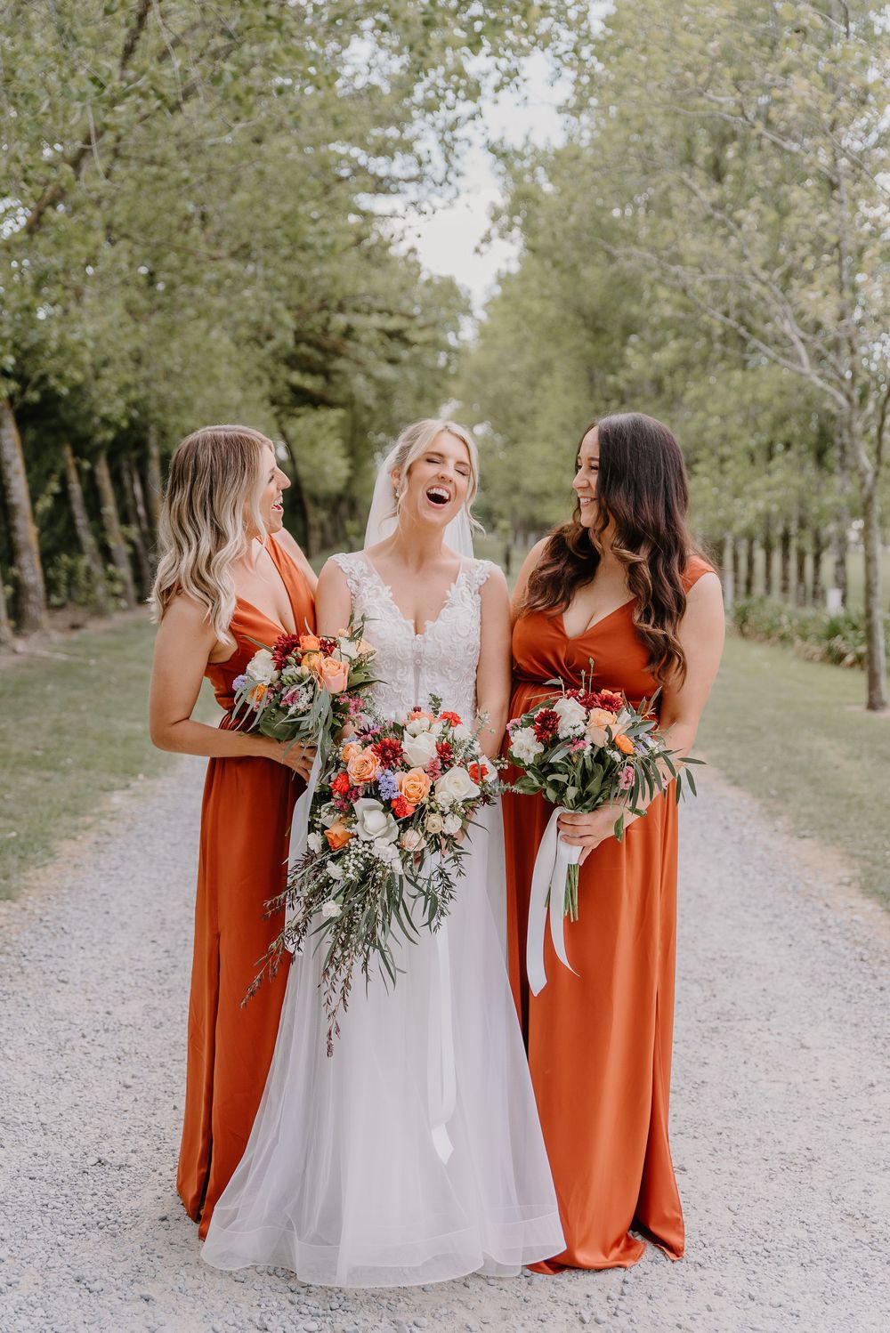A group in orange bridesmaid dresses and white wedding gown walk along a tree-lined gravel path together.