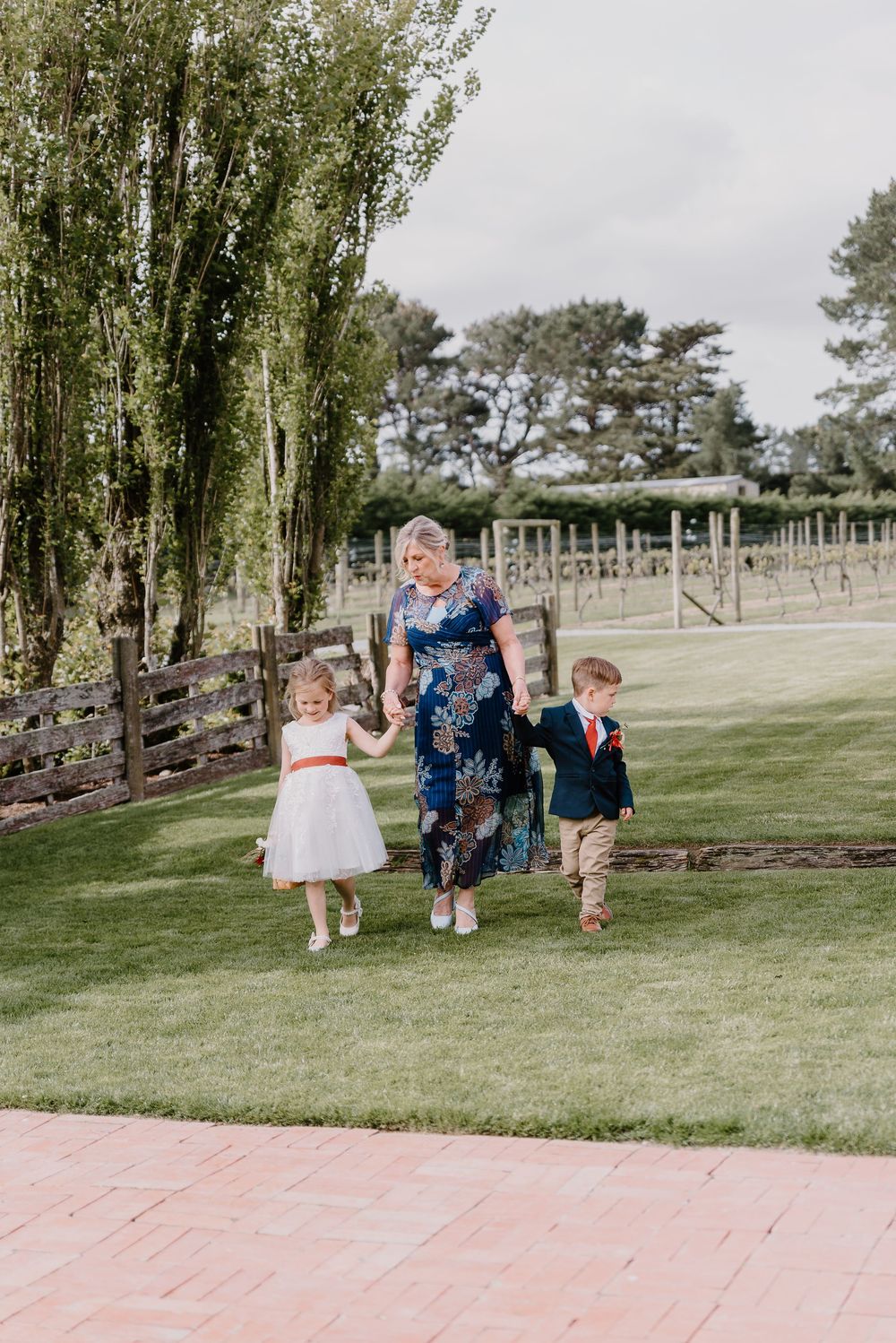 Children in formal attire walk across a green lawn at an outdoor venue with trees in the background.