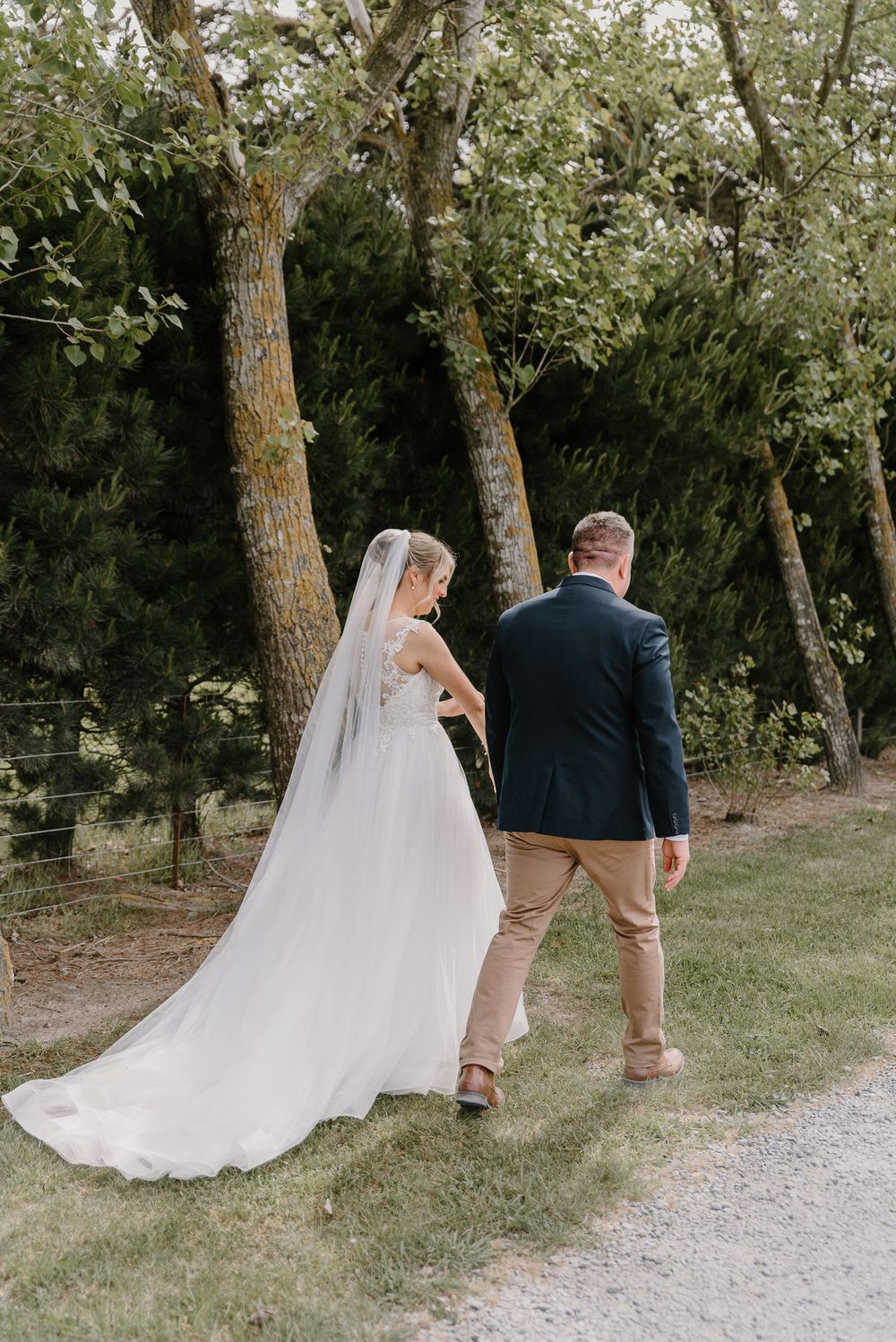 A bride and groom walk down a tree-lined gravel path with her flowing cathedral veil trailing behind them.