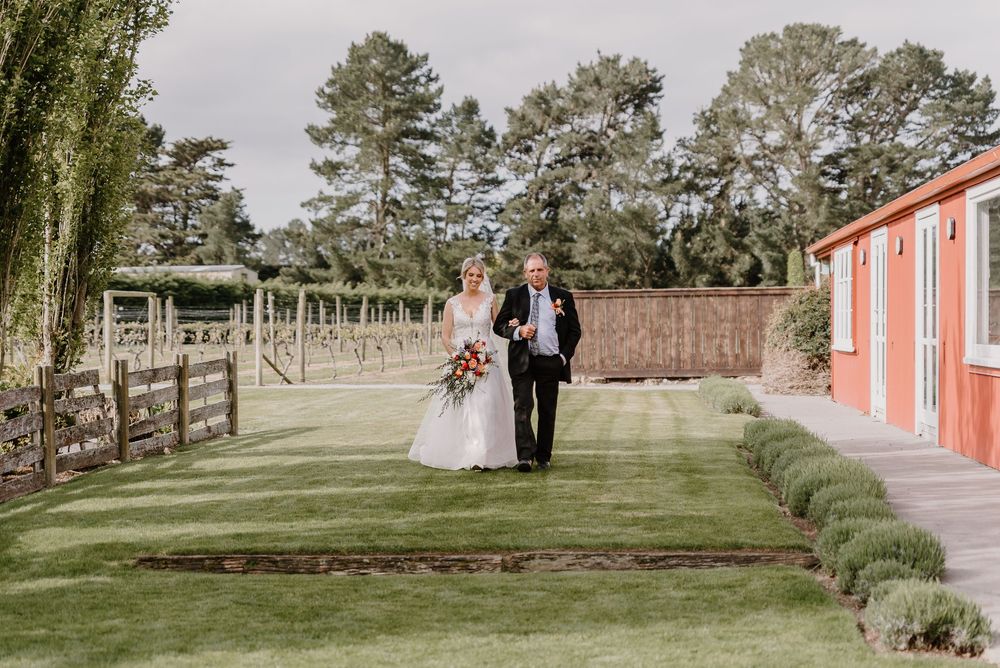 Wedding couple walks through beautifully landscaped garden path lined with coral buildings and pine trees.