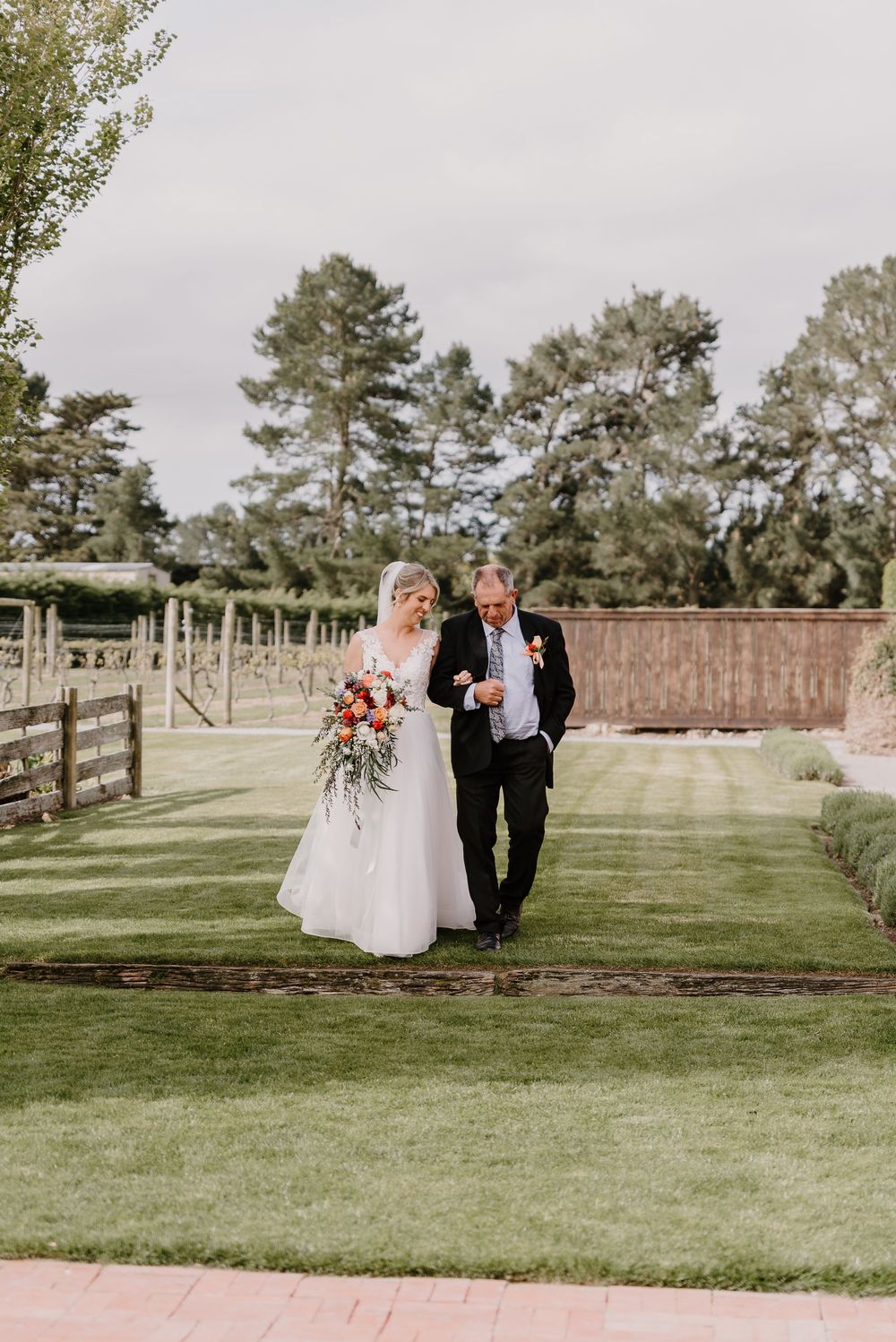 Two figures stroll across a manicured lawn at an outdoor wedding venue with stone walls and pine trees in the background.