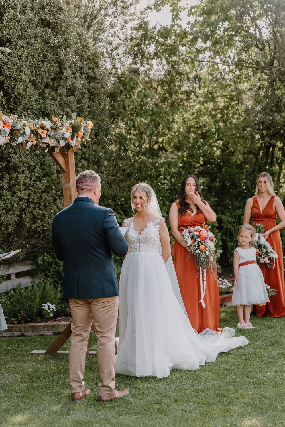 Wedding ceremony takes place in a garden setting with attendants in burnt orange dresses standing beside the bride.