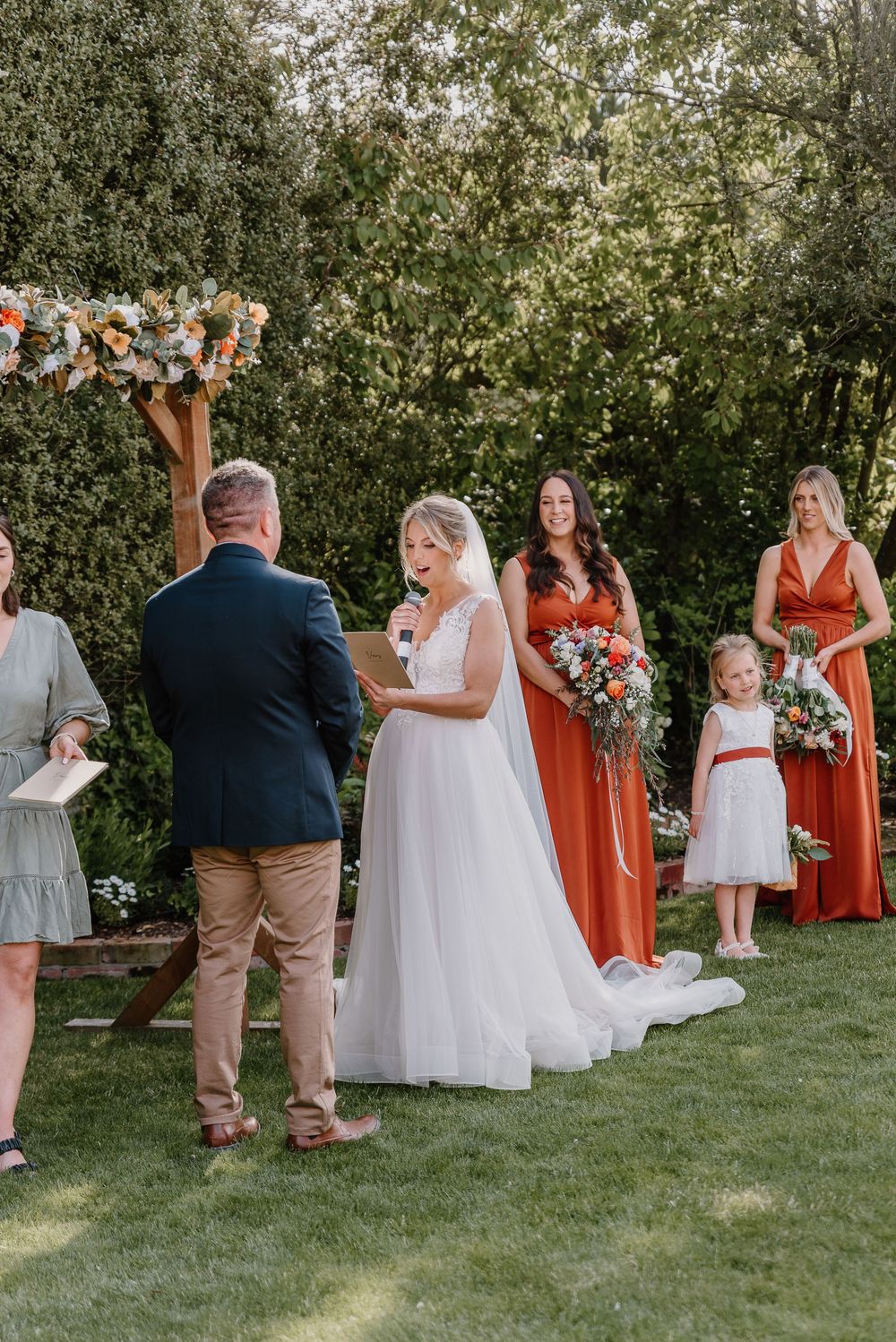 Wedding guests gather on a green lawn with bridesmaids in burnt orange dresses during an outdoor ceremony.