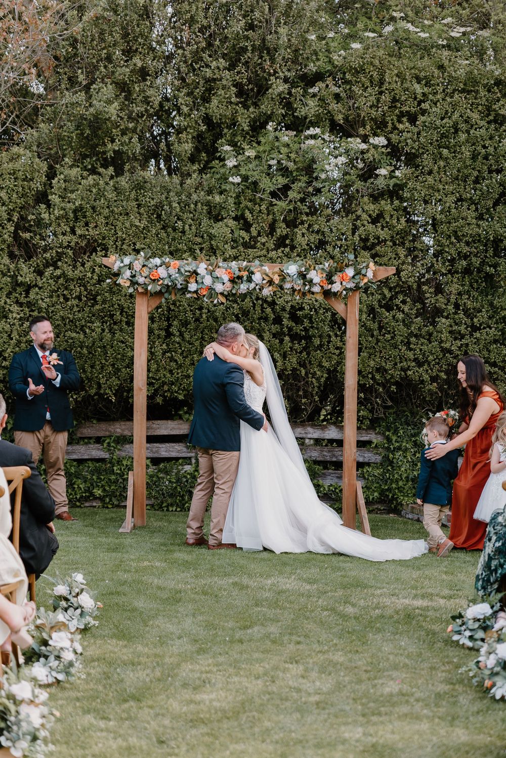 Garden wedding ceremony under wooden arch with floral arrangements and guests gathered on lawn.