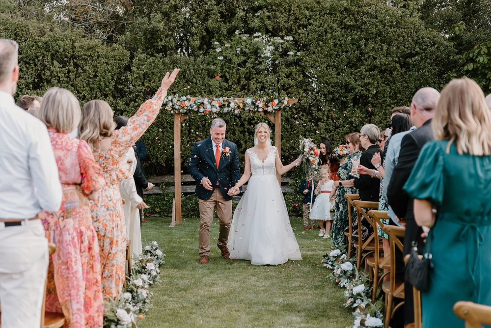 Guests mingle during an outdoor wedding ceremony with a rustic wooden arch and floral decorations in a garden setting.