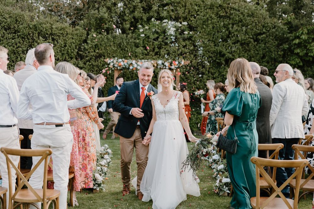 Outdoor garden wedding celebration with guests standing alongside wooden chairs as newlyweds walk down the aisle.