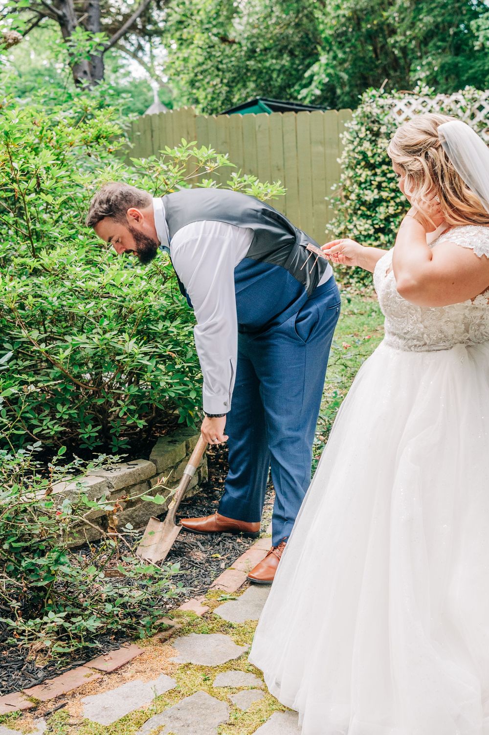 A formal wedding couple stands together in a garden setting next to a pretty stone pathway with green foliage in the background.