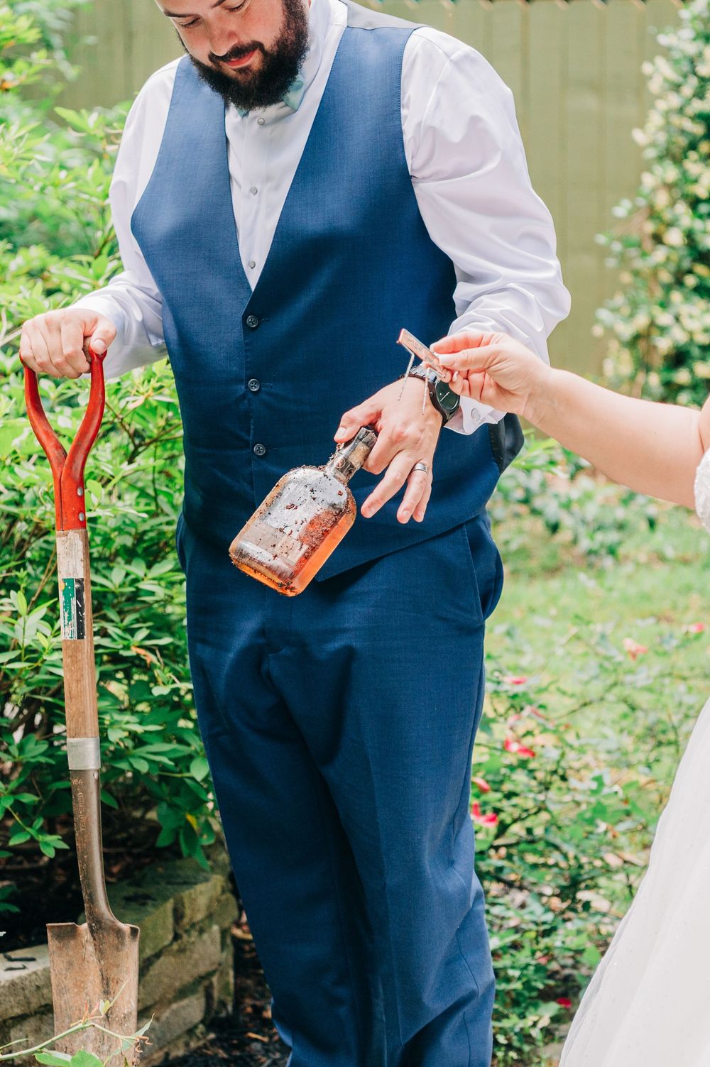 Someone wearing a blue vest and white shirt receives a bottle of whiskey while holding a shovel in a garden setting.