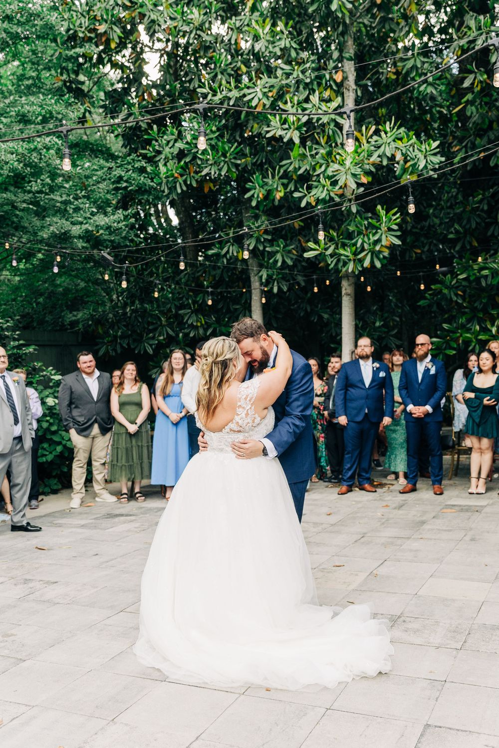 Couple shares their first dance surrounded by wedding guests on an outdoor patio with string lights and greenery.