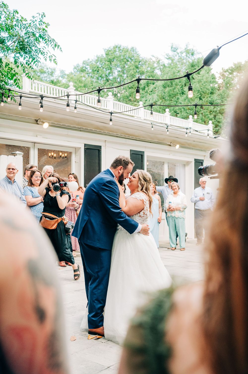 Wedding couple embraces in a tender first kiss while guests look on during outdoor reception.