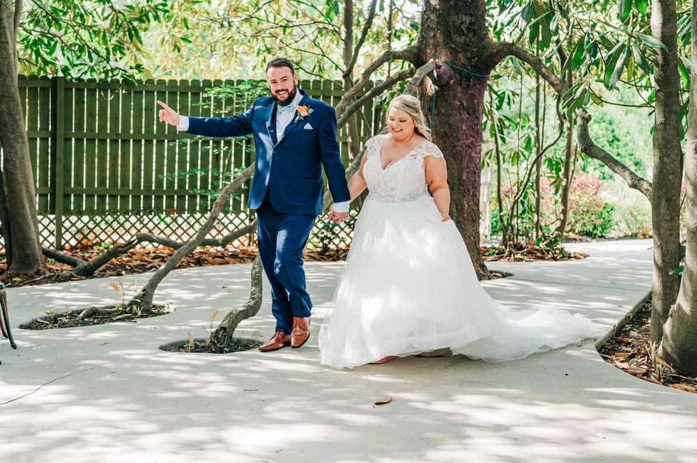 Couple walking and holding hands on a garden path surrounded by trees, the bride in white gown and groom in navy suit.