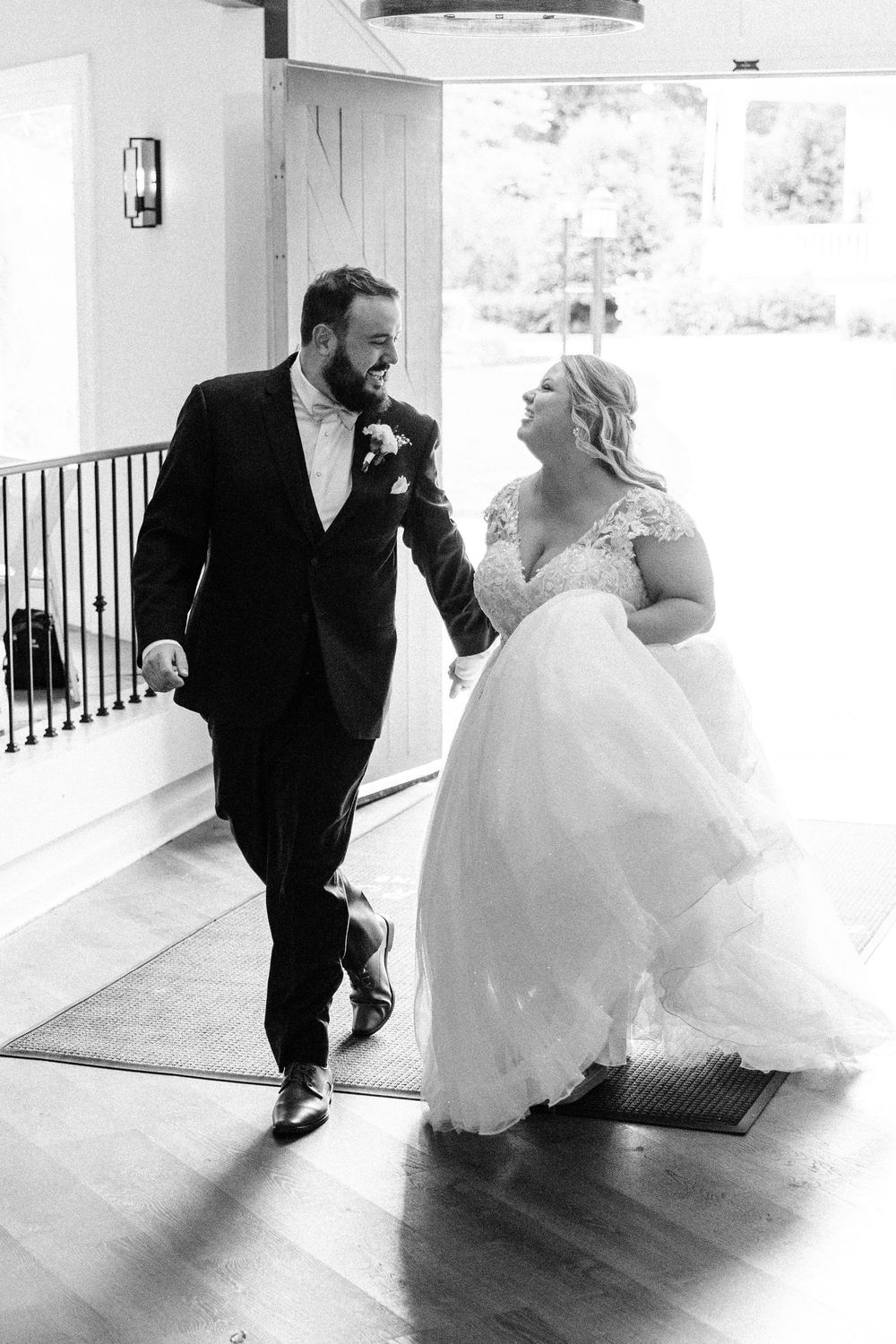 A black and white wedding photograph capturing a joyful moment between a couple in formal attire walking indoors.