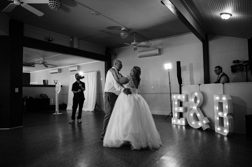 Black and white photo of a couple dancing at a wedding reception with illuminated letters and others in the background.
