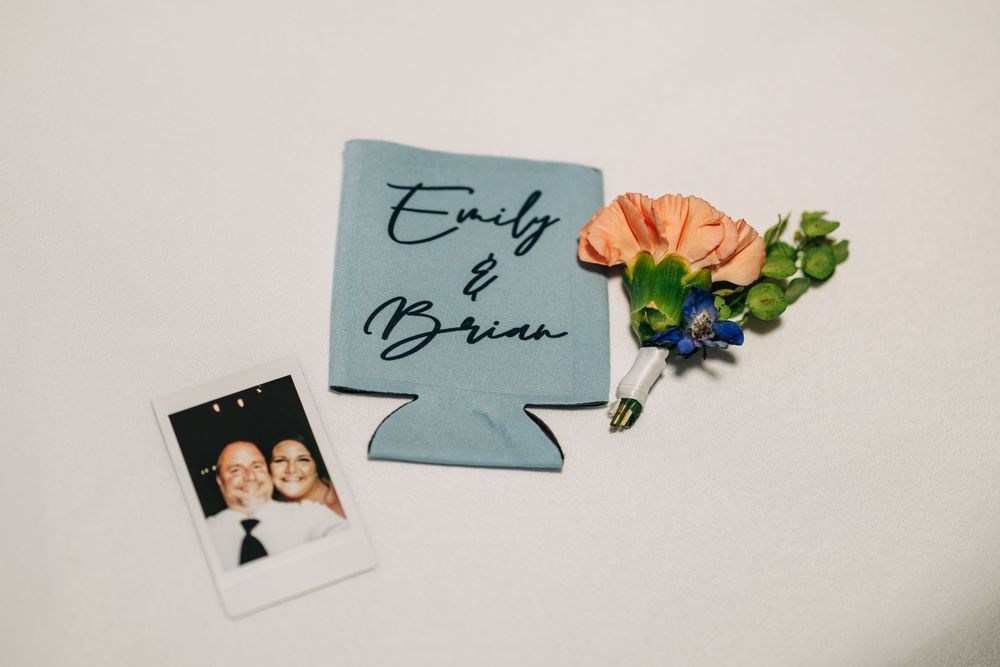 A blue wedding koozie with names, a coral boutonniere with greenery, and a polaroid photo arranged on a light surface.