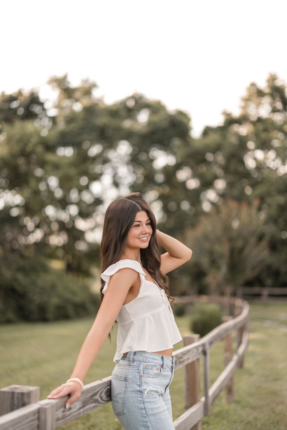 A woman in a white sleeveless top and jeans leans against a wooden fence in a sunny outdoor setting with trees.