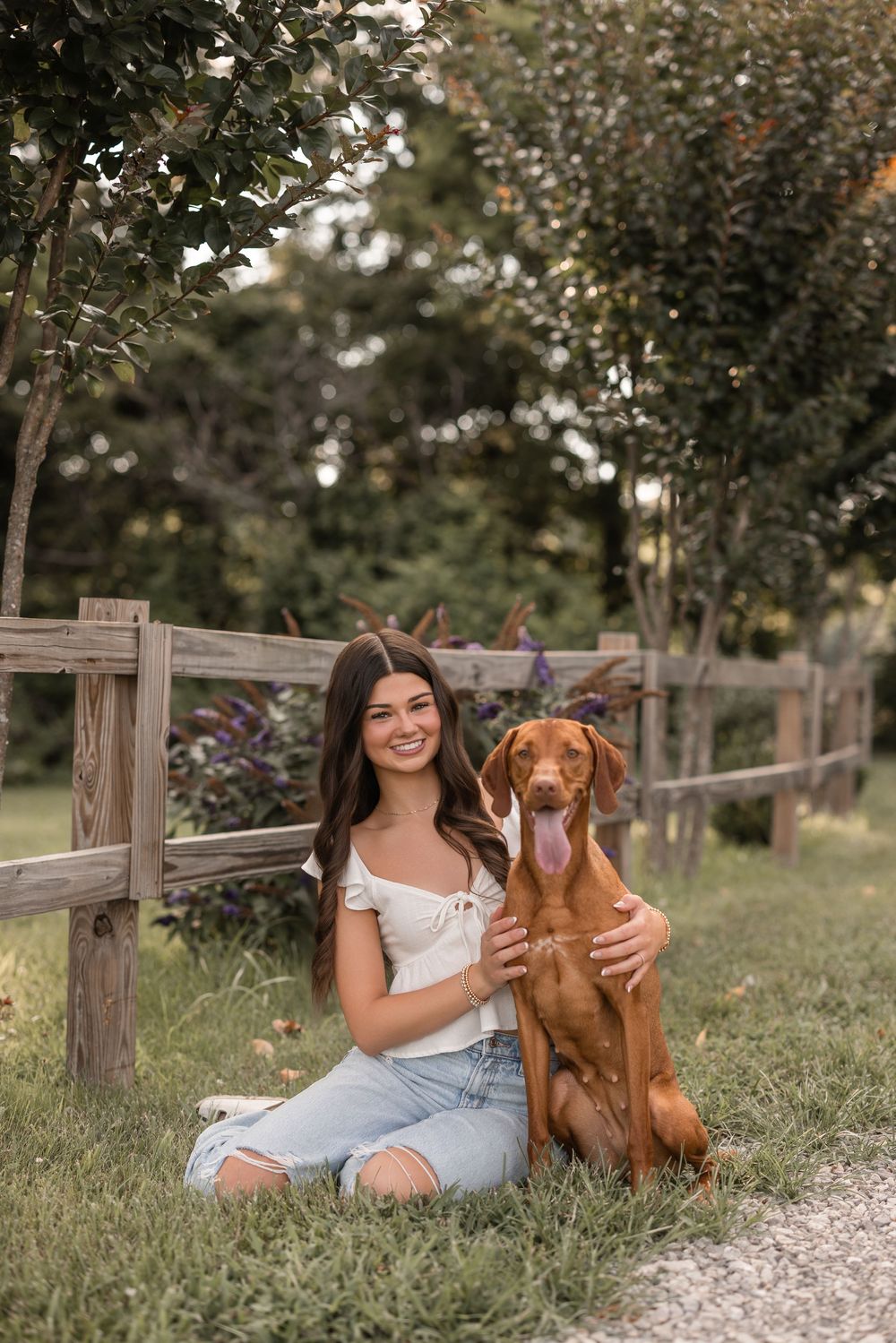 A radiant figure in a white top and denim shorts sits by a wooden fence with their happy Vizsla dog companion.