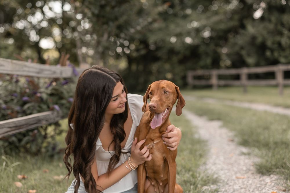 A Vizsla dog and companion share a tender moment together on a peaceful outdoor path surrounded by nature.