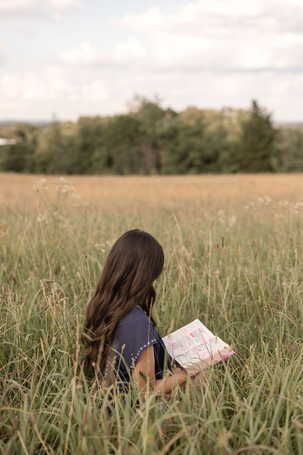 A reader with long dark hair sits peacefully in a tall grass meadow holding an open book on a tranquil afternoon.