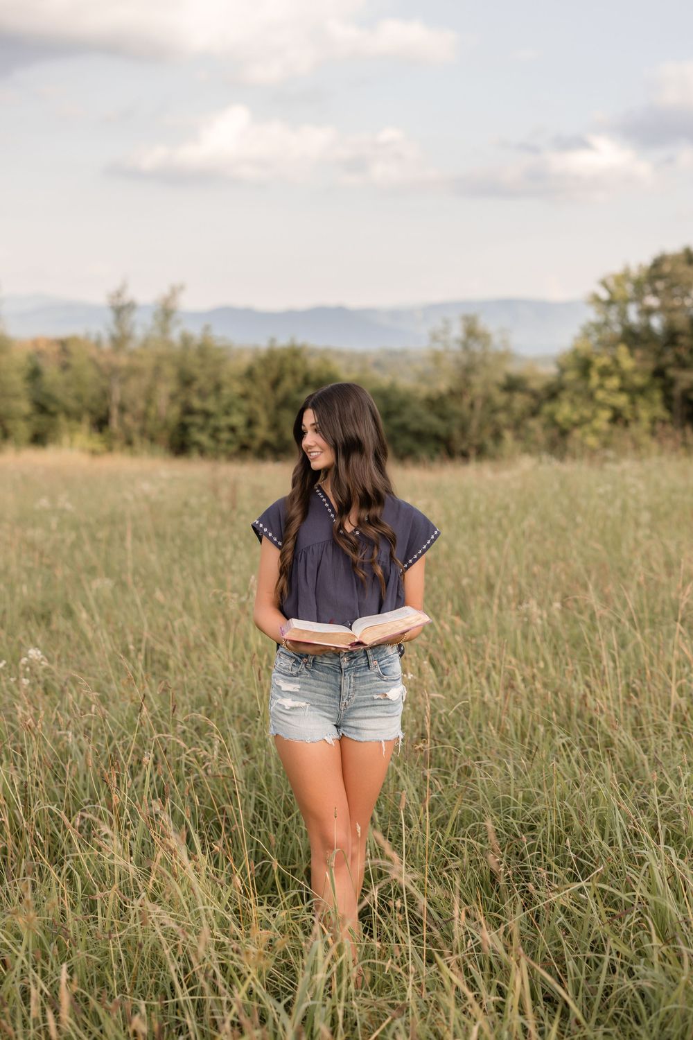 A woman in denim shorts and navy shirt stands in a sun-lit meadow with mountains visible in the background.