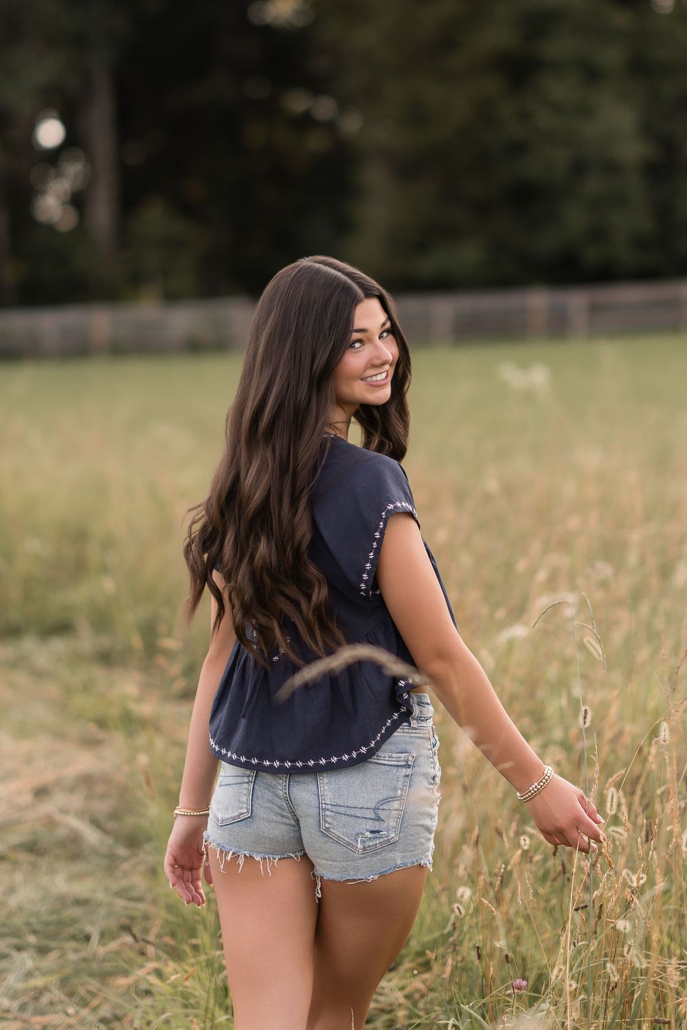 A person wearing a navy top and denim shorts stands in a sunlit meadow with tall grass.
