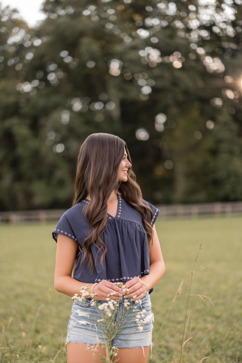 Woman in navy top and denim shorts holding wildflowers in a sunny countryside field at dusk.