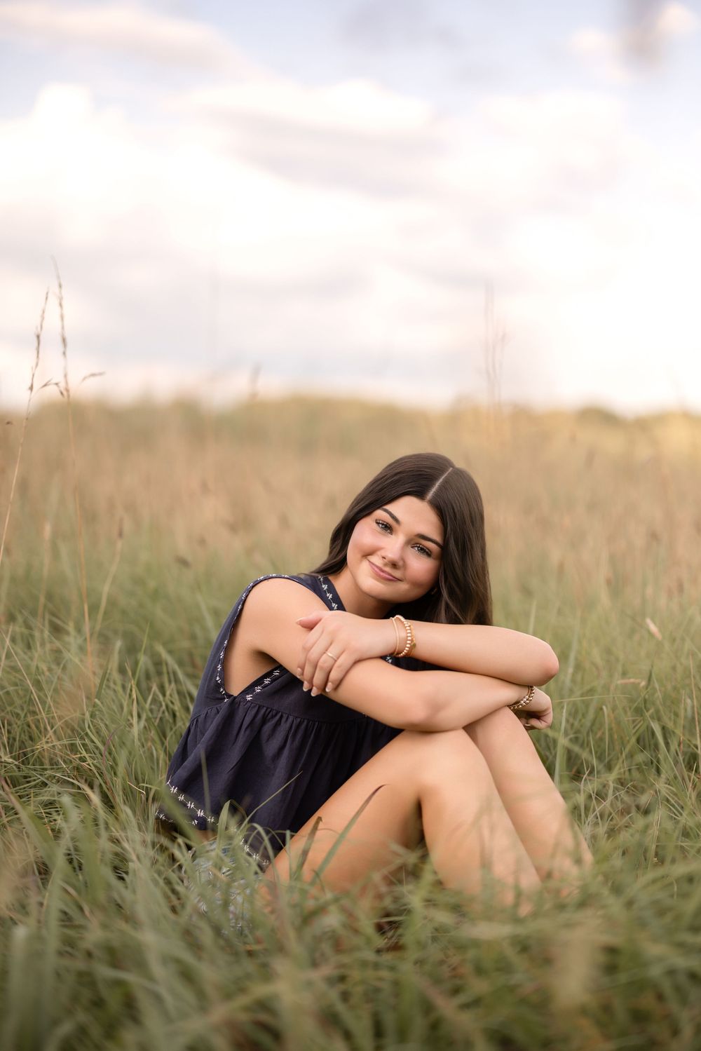 A tranquil outdoor portrait of a person in a navy dress sitting in tall grass during golden hour.