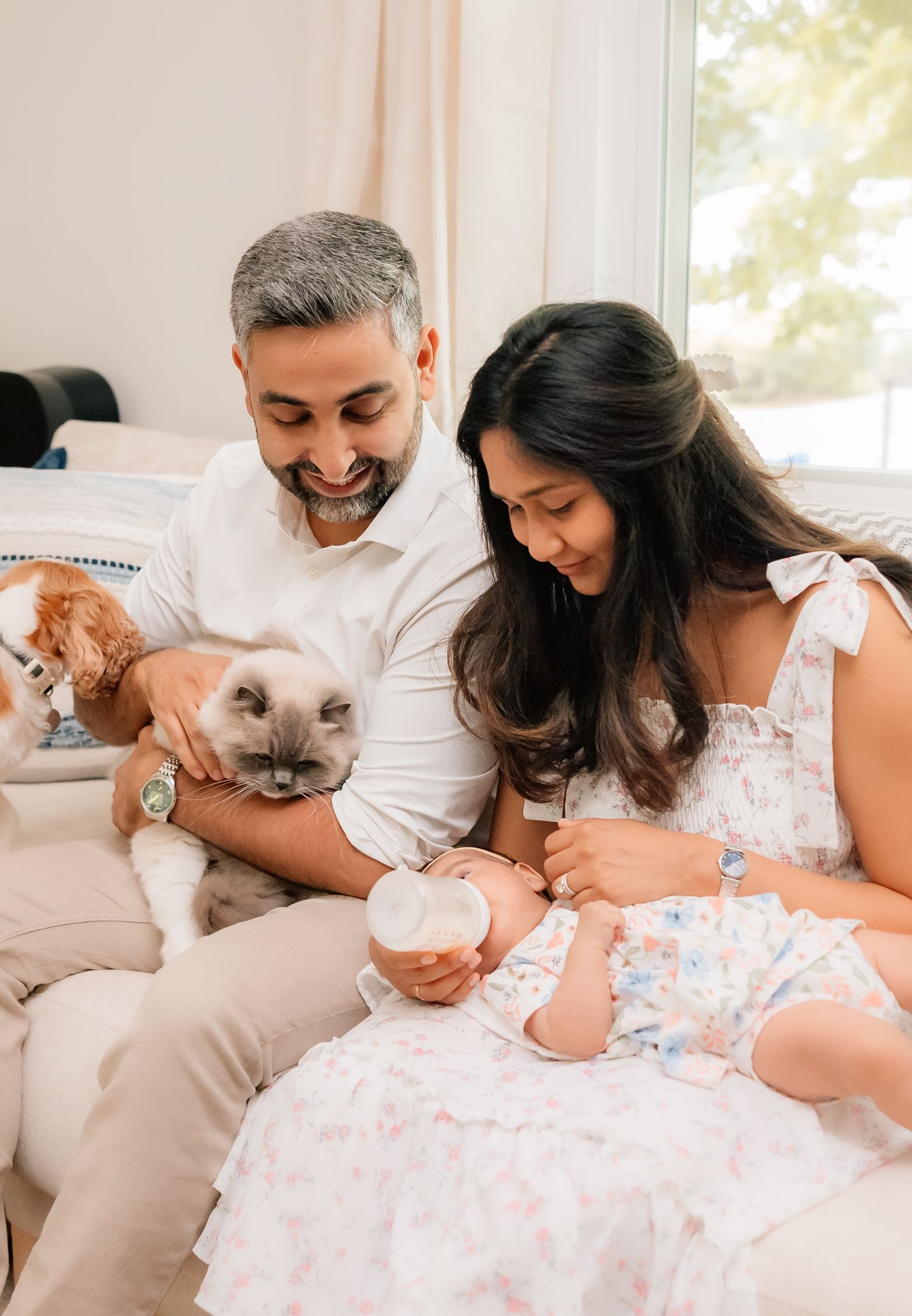 A couple snuggles close to their baby while lying on a white couch with their Siamese cat joining the family moment.