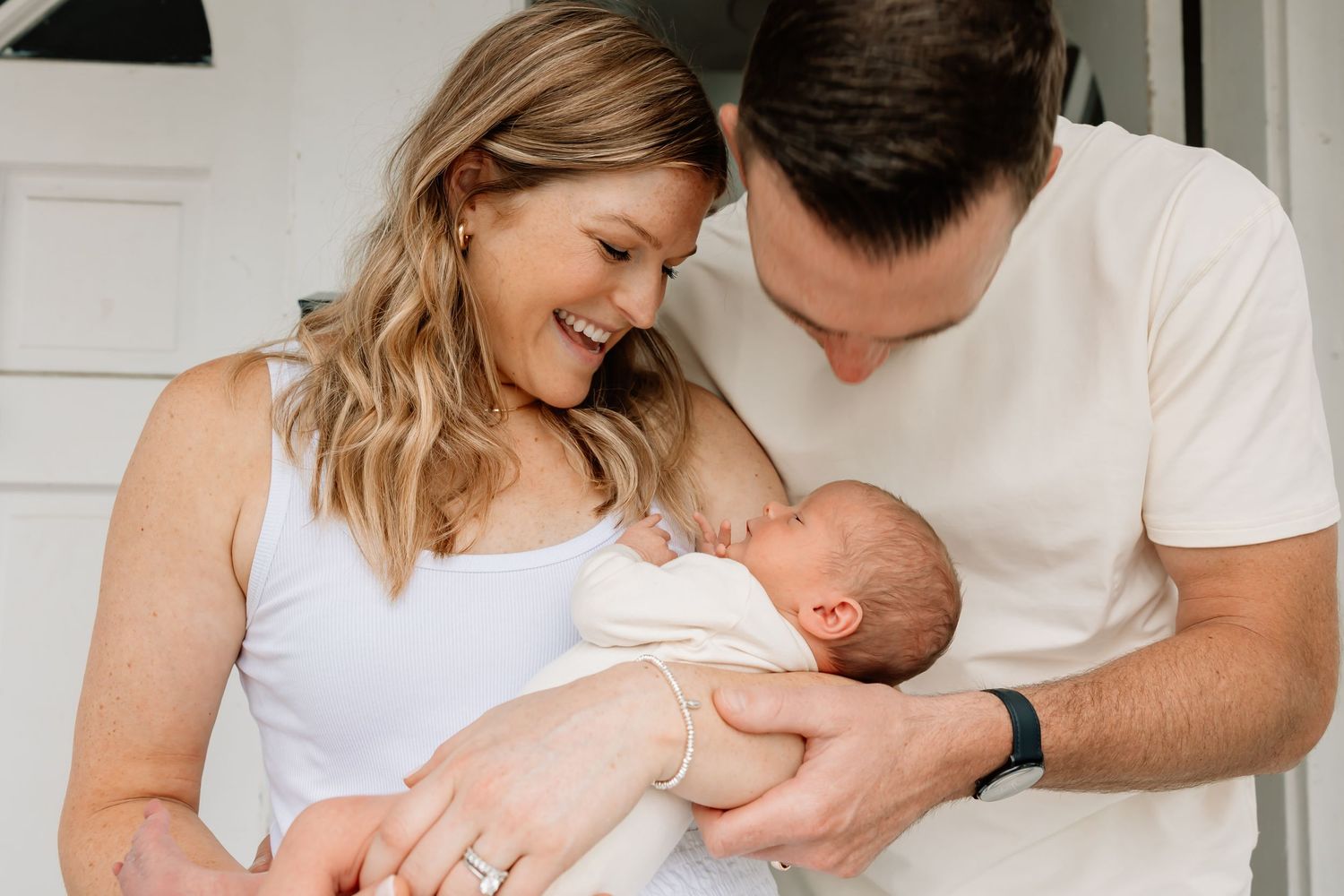 Parents lovingly holding and gazing at their newborn baby while wearing white casual attire at home.