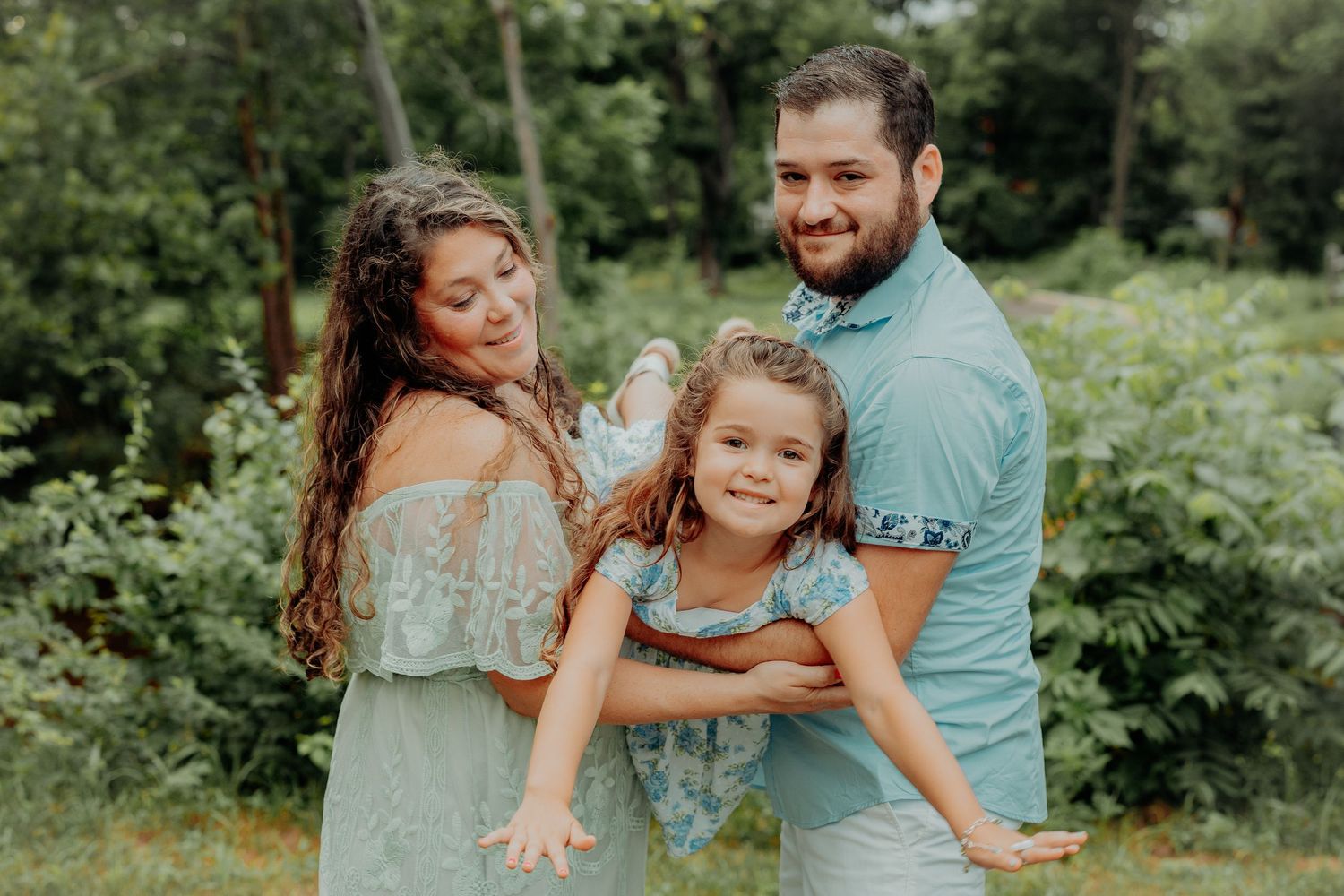 Family wearing coordinated sage green and white outfits enjoys playful moments together in a lush garden setting.