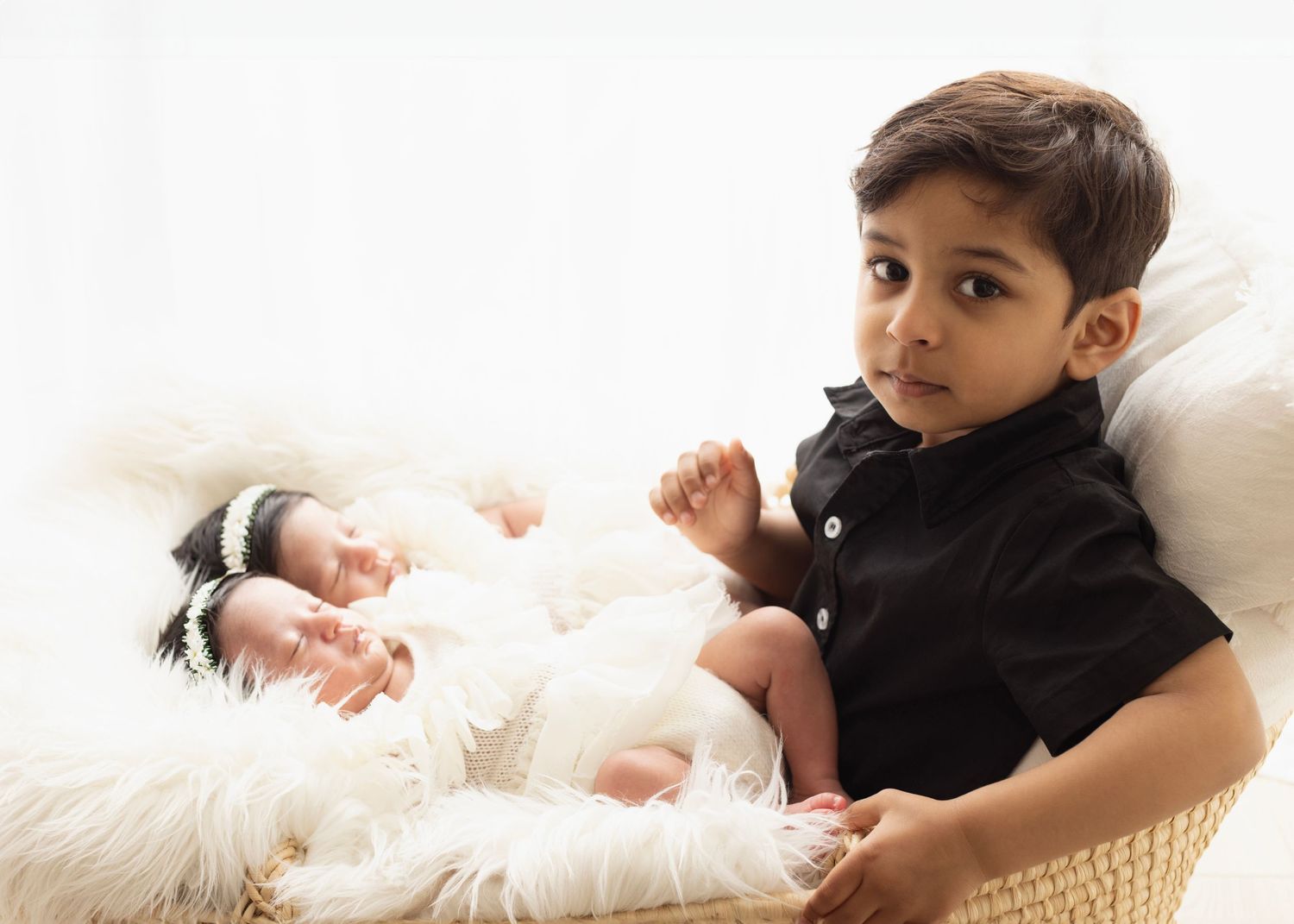 Big brother holds his baby newborn sisters on his lap. He looks to the camera. Such a small boy but yet a big brother. Surrey.