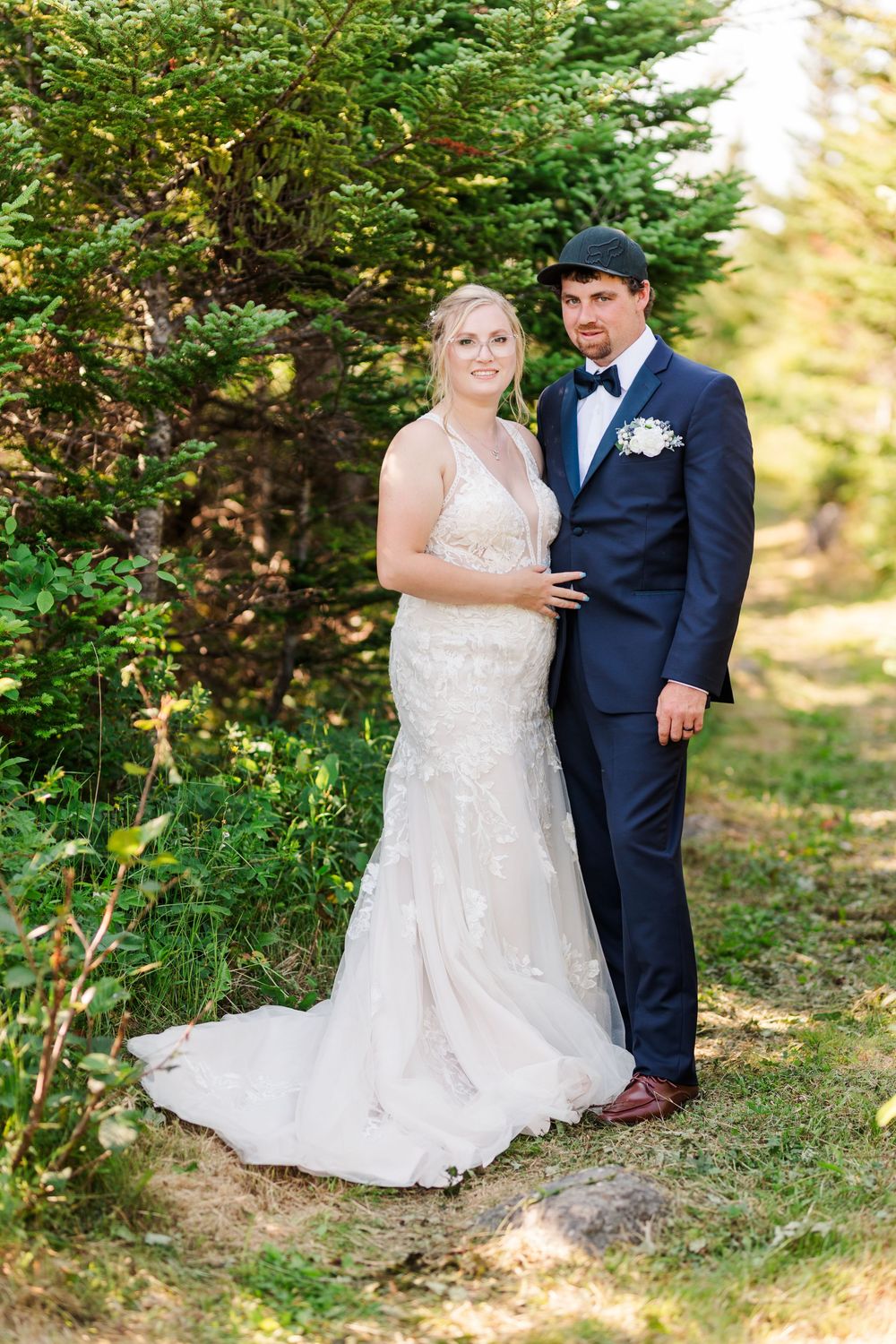 A bride and groom pose together outdoors among evergreen trees in a natural woodland setting on their wedding day.