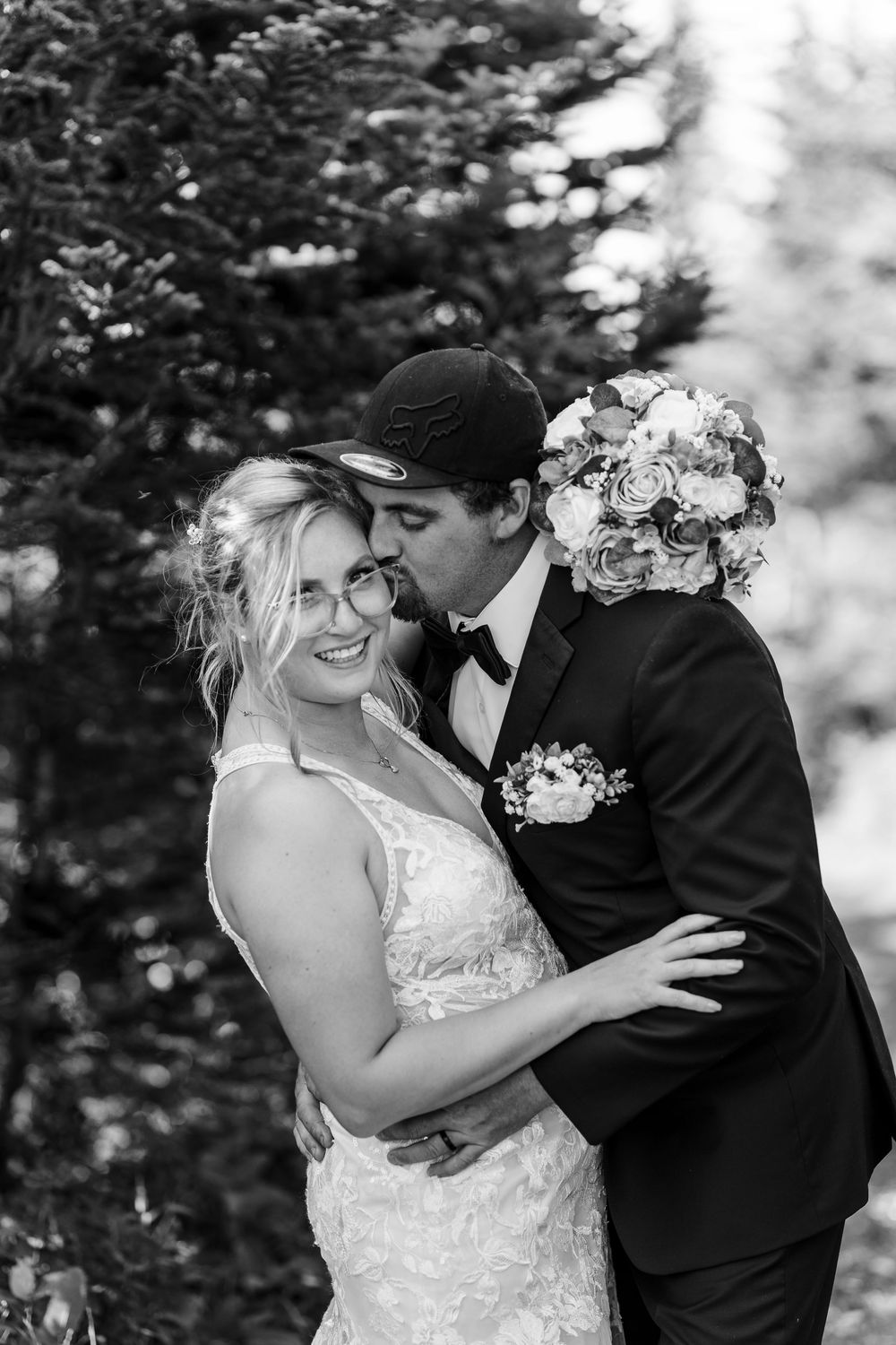 A black and white wedding photograph showing a tender moment between a couple outdoors with a floral bouquet.
