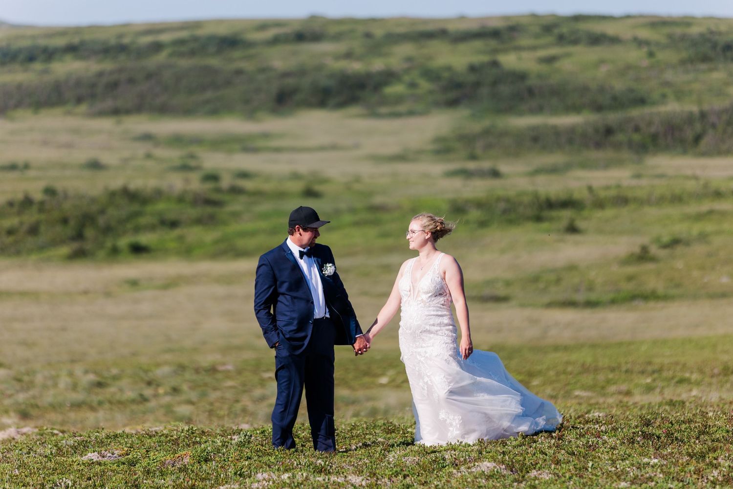 A newly married couple in formal wedding attire enjoys a quiet moment together in a rolling green field on their special day.