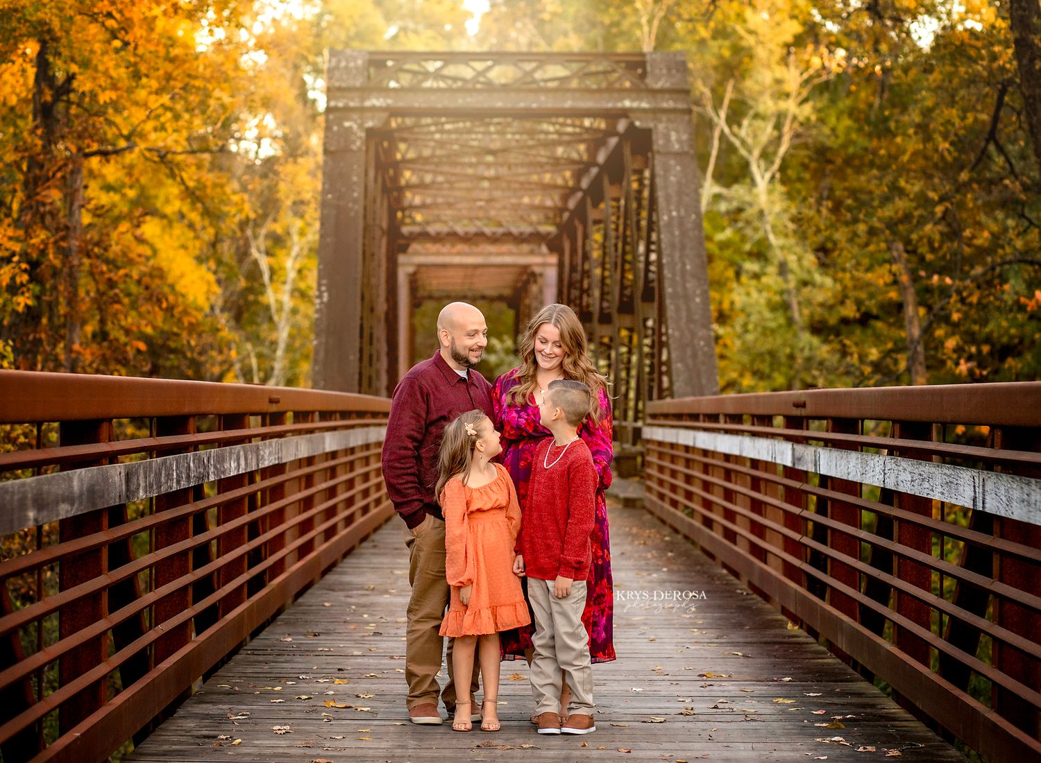 A couple walks along a wooden bridge surrounded by vibrant autumn foliage in a romantic outdoor setting.