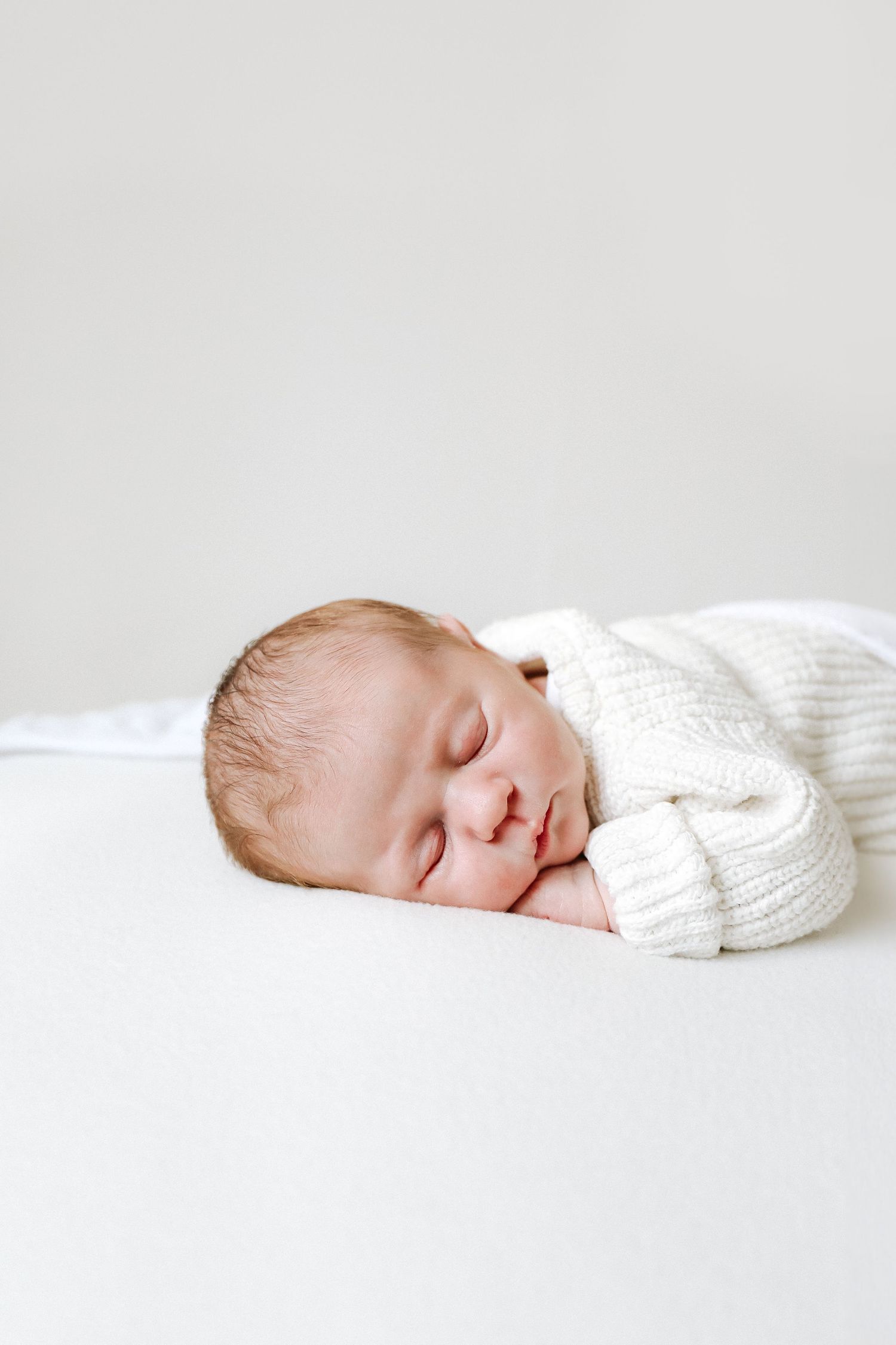 A peaceful sleeping baby wearing a soft white knitted sweater lies on white bedding.