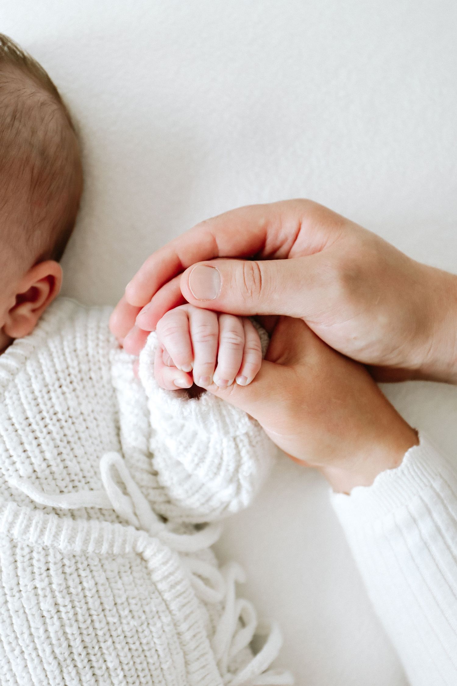 A tiny baby's hand is tenderly held in an adult's grasp while wearing a white knitted sweater.