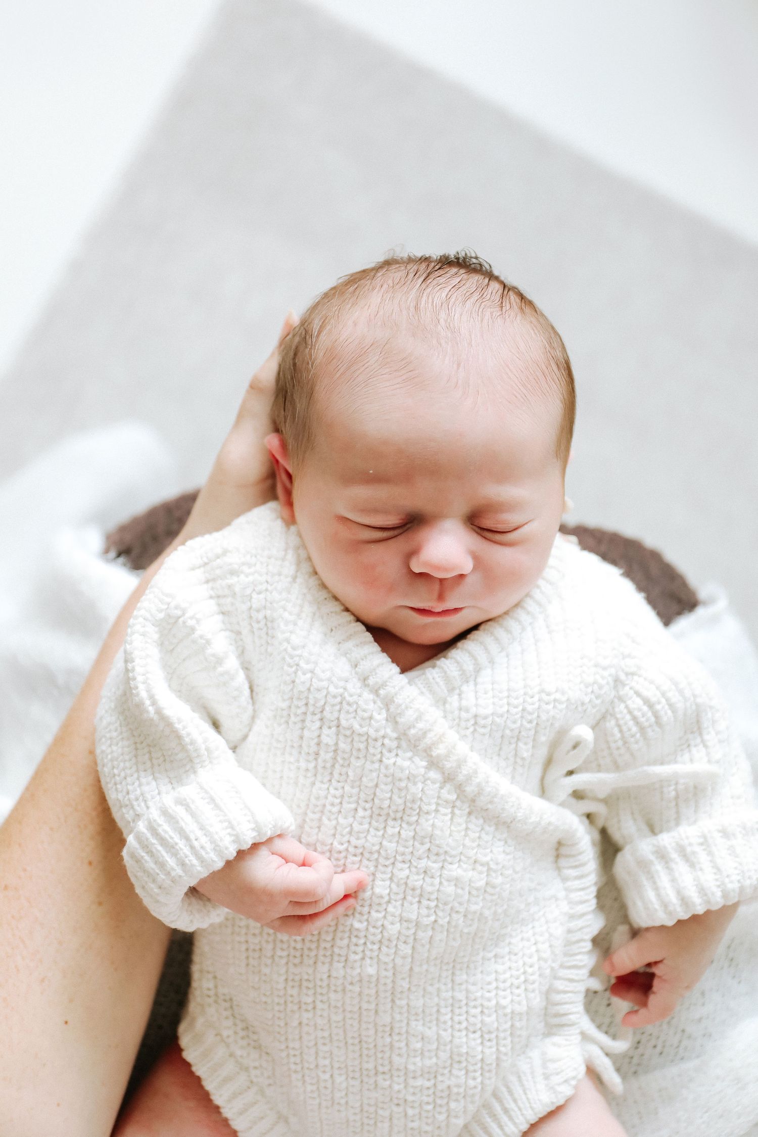A sleeping newborn baby wearing a soft white knit sweater lying on a light-coloured blanket.