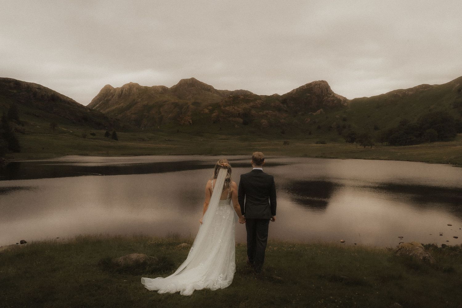 A couple stands at a tranquil lakeside with mountains in the background during a moody sunset intimate wedding at Blea Tarn.
