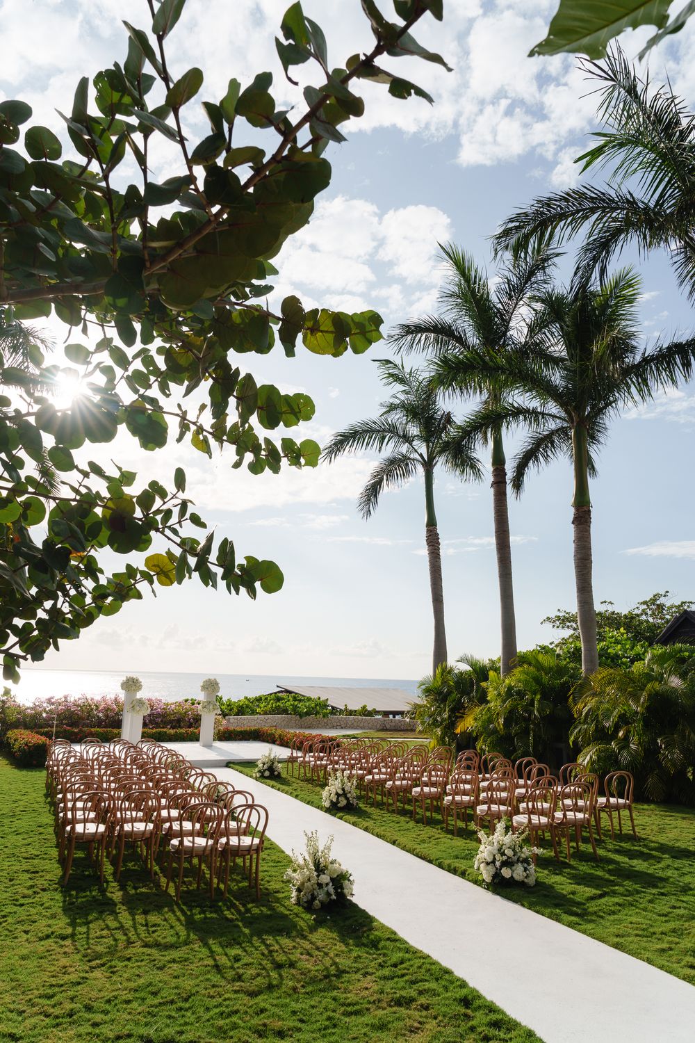 Outdoor wedding ceremony setup with rows of wooden chairs along white aisle runner framed by tall palm trees and ocean view.