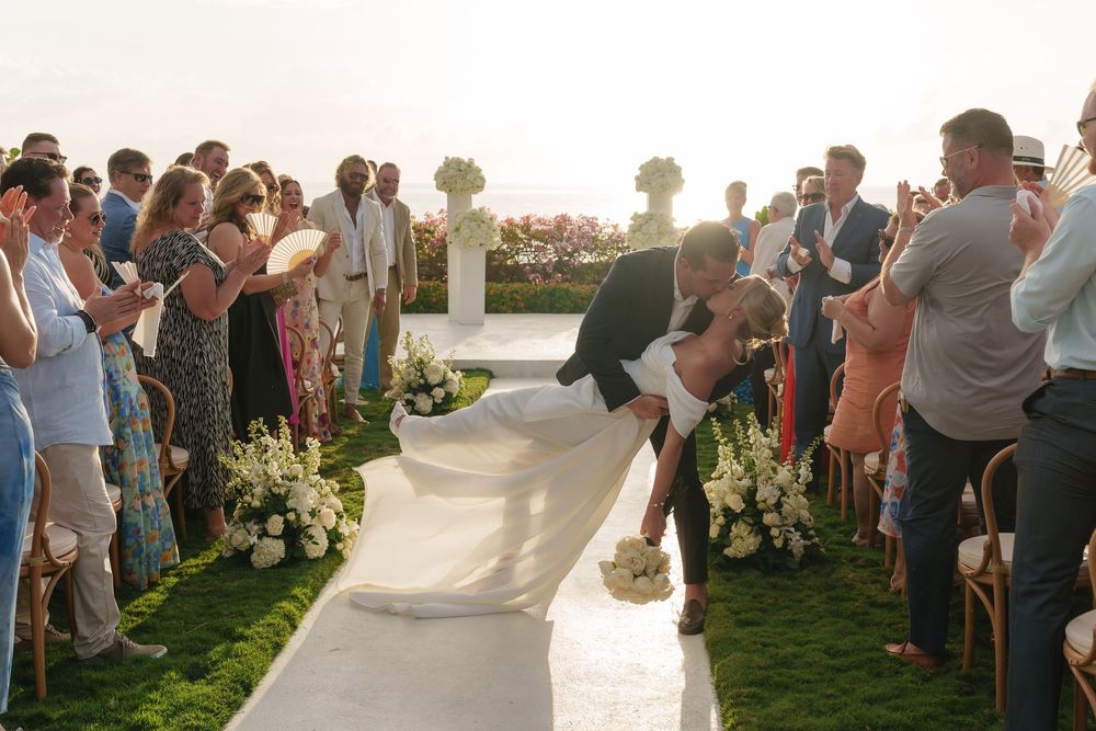 Wedding guests gather at an outdoor ceremony by the water, with a white tent and chairs arranged on the grass.
