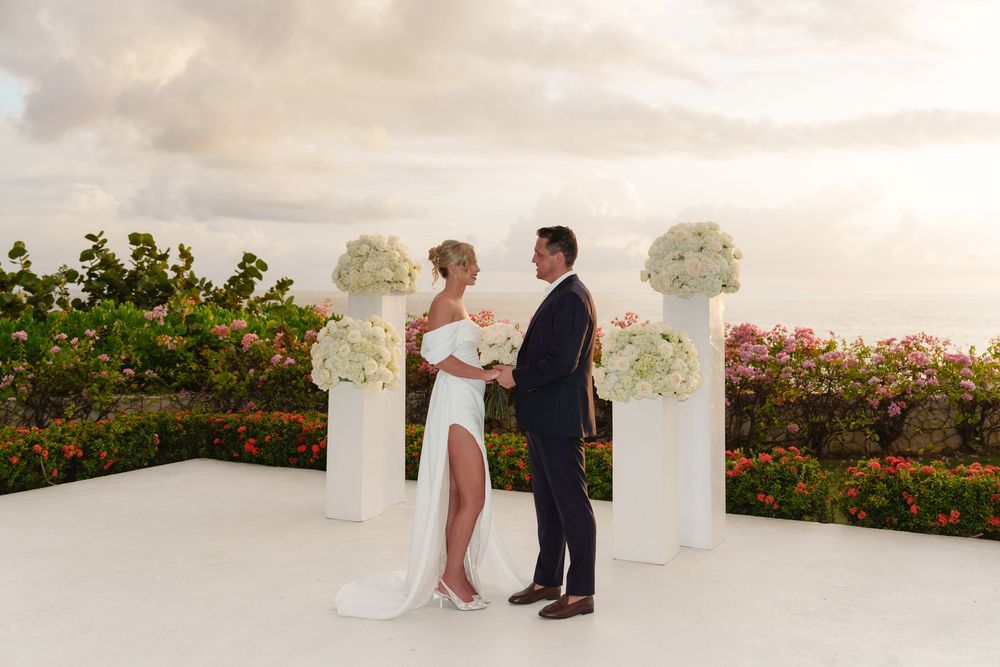 A romantic wedding ceremony takes place at sunset on a white platform surrounded by floral arrangements and gardens.