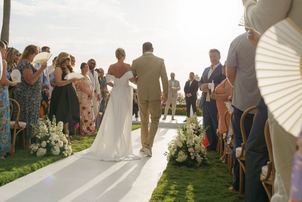 A couple walks down an outdoor wedding aisle lined with guests and floral arrangements during a sunny ceremony.