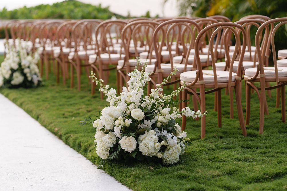 Elegant wooden bentwood chairs lined up on grass with lush white floral arrangements for outdoor wedding ceremony.
