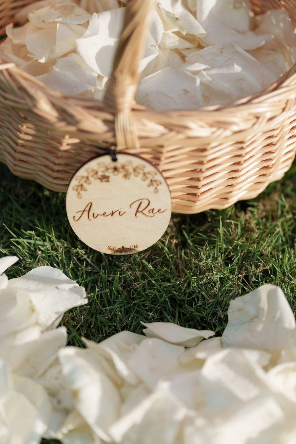 A wicker basket with white rose petals and a circular wooden name tag labeled 'Aerin Rae' rests on grass.